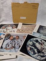 Photo of original vintage brown mailer envelope with visible address label and multiple Apollo mission photographic prints spread out beneath it including astronaut group portraits and lunar surface photos.