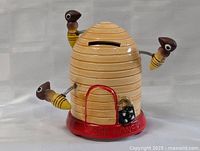 Front angled view of the ceramic beehive-shaped honey bank with three bees on springs, coin slot on top, door at front, and red base with raised inscription 'BILLY BEE HONEY'.