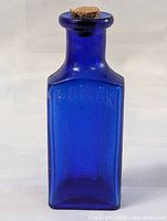 Front view of the blue glass poison bottle with the cork stopper in place, showing the embossed word 'POISON'.