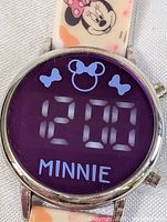 Close-up photo of the round digital watch face showing the time 12:00 with Minnie Mouse icons and the word 'MINNIE' in blue on purple background.