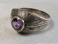 Front and angled view of the sterling silver ring with a faceted oval amethyst gemstone set in the middle. Shows the textured sunburst design on the band.