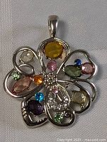 Close-up of flower-shaped pendant with multiple colored stones in silver-tone setting on white fabric background.