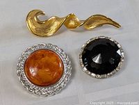 Three pin brooches: gold-tone abstract wave shape, round amber cabochon in silver-tone twisted frame, round black faceted stone in silver-tone setting.