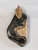 Front view of an irregular polished stone pendant with distinct black and beige swirled pattern, attached to a gold-tone bail shaped like a leaf.