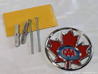 Photo showing the Canadian Automobile Association badge alongside its three mounting bolts and a yellow envelope mailer.