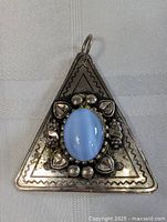 Front close-up image of triangular silver pendant with embossed heart details and central light blue cabochon stone.