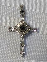 Front view of silver-tone stylized cross pendant on white textured fabric showing central cabochon stone and metal details.