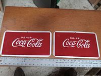 Photo showing 2 rectangular Coca-Cola metal signs side by side on a wooden surface with ruler nearby measuring size of signs.