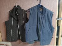 Two men's sleeveless vests hanging on hangers against wood wall, one dark quilted, one blue with fleece lining.