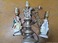 Photo showing two porcelain lamps with couple figurines and one brass painted lamp with cherubs together on a wooden surface