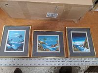 Three framed coastal landscape paintings by Manolete Pastol with a ruler for scale, showing their size and details.