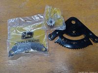 Photo showing three John Deere parts: one metal gear packaged in branded plastic, a smaller metal gear still packaged, and a black metal mounting bracket with a gear rack and mounting holes.