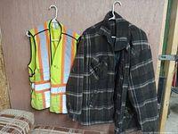 Construction safety vest in neon yellow with bright orange reflective stripes and men's black-gray plaid spring coat hanging on wall