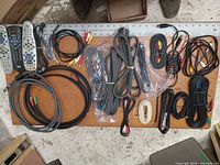 Top-down view of multiple coiled cables and remote controls on a wooden floor, various AV cables identifiable by connectors such as RCA and component video colors, some cables plastic wrapped.