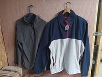 Pair of men’s sweaters hanging against a wooden panel background. Left is a plain grey quarter-zip pullover. Right is a navy and light grey fleece with maroon accents.