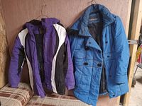 Photo showing both coats together hanging on a wall - purple ski jacket and blue Nevada quilted coat.