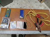 Photo showing welding glass lens tins, rebar wire ties in plastic bag, jump cables, small metal parts and fasteners on wooden table.