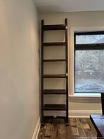 Full height bookshelf positioned against wall near window, showing six dark stained wooden shelves supported by black steel frame with bolts.
