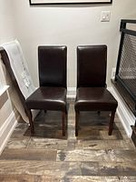 Pair of brown leather dining chairs with straight backs and wooden legs on a hardwood floor.