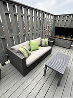 Dark brown wicker sofa on deck with beige seat cushion and three green throw pillows, grey coffee table in front