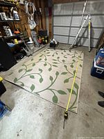 Photo showing a large outdoor rug laid flat on a concrete floor with a tape measure along one side indicating its size. The rug has a beige background and green leaf and vine design.