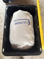 Photo of white storage bag with AeroBed logo containing queen size inflatable bed inside a black plastic storage bin.