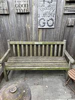 Full view of teak bench against wooden fence showing slatted backrest, seat and armrests with weathered finish.