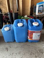 Photo of three blue plastic water jugs of different sizes with white screw-top lids, placed on a wooden floor against a wall.