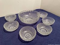 Seven clear textured glass bowls arranged on purple cloth, one is larger with floral textured design inside and six smaller matching bowls.