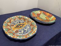 Two Mexican Talavera ceramic plates on a dark blue cloth surface showing their full design and shape.