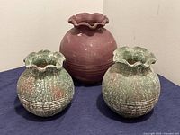 Three clay vases: two smaller green and brown vases with textured scalloped rims, one larger reddish-brown vase with textured scalloped rim; arranged on a dark fabric fabric surface against a plain wall.