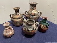 Five decorative Mexican folk art style metal vases showing front and side views on a blue cloth background.