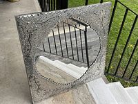 Square silver pressed tin framed wall mirror on steps, showing front reflective surface with ornate floral and dotted embossed pattern.