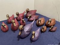 Full set of 15 decorative clay ducks arranged on a blue cloth, showing different colors including purple, red, and brown with shiny gold metallic beaks and textured feather details.