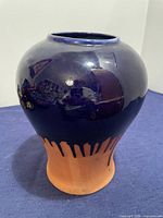 Front view of a bulbous Mexican clay pot with a glossy dark blue glaze dripping down to a natural terra cotta base, placed on a purple surface indoors.