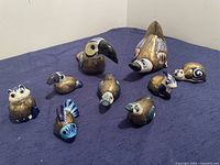 Image shows full set of nine Talavera ceramic animal figurines arranged on a blue surface including a toucan, raccoon, owl, bird, snail, and other small animals, all hand-painted with metal details.