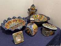 Full set of hand-painted Talavera ceramic items displayed on a dark cloth, showing colorful floral and geometric patterns on each piece, including serving dish, basin, bottle, frame, jewelry case, and wall decoration.