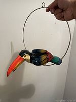 Front and side view of first ceramic toucan painted in bright orange, blue, black, green and red mounted on metal ring for hanging
