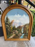 Front view of the painting showing the mountain landscape with waterfall, hut, trees, and river within a curved wooden frame.