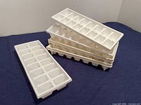 Five white plastic ice cube trays stacked, showing their shape and size with rectangular compartments for ice molding.