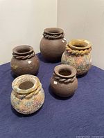 Five rustic Mexican clay pots arranged on a blue cloth surface, showing variety in size and coloration, with twisted rope detail on each pot's neck.