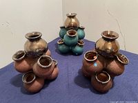 Group of three decorative multi-neck vases in bronze, turquoise, and copper colors, displayed on a cloth surface.
