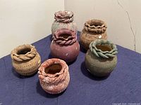 Set of six small Mexican pottery vases in assorted earth tone colors, arranged on a dark cloth. Each vase has a twisted rim and carved patterned surface.