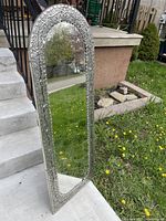 Full view of the mirror leaning outdoors, showing the full-length rectangular shape with arched top and detailed silver embossed floral frame.