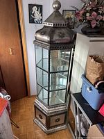 Full view of the vintage hexagonal display cabinet showing glass and metal construction with decorative motifs and a tall finial.