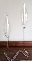 Pair of clear glass teardrop oil lamps with candle wicks inside on uniquely shaped clear glass stands shown against white wall and wood baseboard.