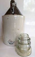 Stoneware crock jug and glass insulator showing both items from front and side to highlight details such as handle and emblem.