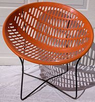 Orange molded plastic circular chair seat with perforated pattern on black metal frame, shown outdoors on white cloth backdrop.