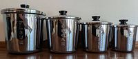 Group photo of the four Everedy stainless steel canisters labeled Flour, Sugar, Coffee, and Tea with black lids placed on wooden surface.