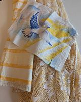 Three different linen tea towels laid out: a 1984 calendar with bird and floral design, a yellow and white striped towel, and a beige floral lace towel.